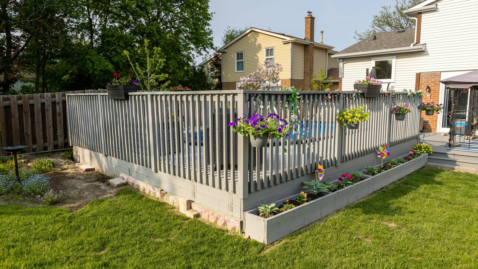 A gray wooden fence around a back yard pool deck with raised planter beds of hostas and annuals along its base, lawn and neighboring houses behind